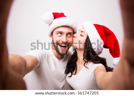 christmas, holidays, technology and people concept - happy couple in santa hats taking selfie picture from hand on white background. She kiss him.