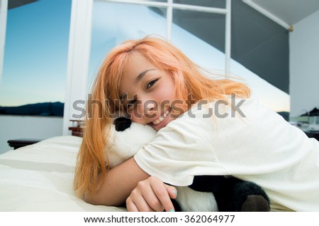 Pretty young woman lying comfortably on white bed and hugging stuffed panda animal with large windows in background.