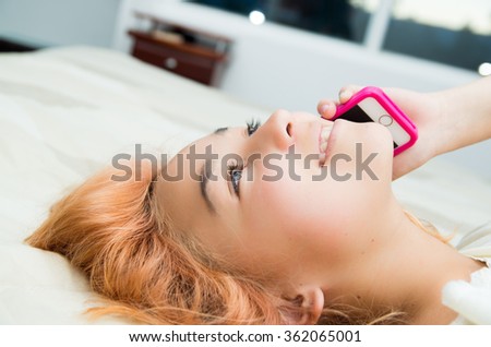 Pretty young woman lying comfortably on bed and using pink mobile phone with large windows background.