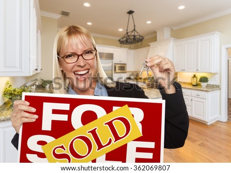 Happy Young Woman Holding Sold For Sale Real Estate Sign and Keys Inside Beautiful Custom Kitchen.
