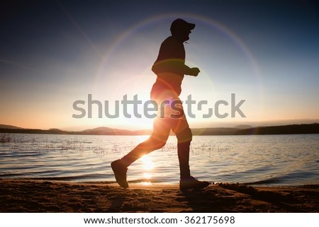 Running man in Sun rays on beach. Sportsman in baseball cap, jogging during the sunrise above sea level. Strong flare effect.