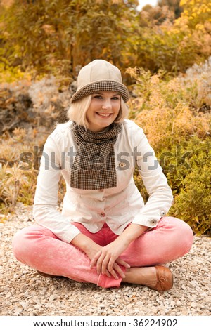 Picture of a young happy smiling blond girl in a hat sitting in the autumn park near the lake