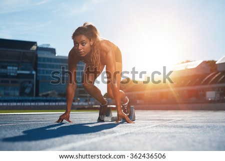 Confident female athlete in starting position ready for running. Young woman about to start a sprint looking away with bright sunlight from behind. Royalty-Free Stock Photo #362436506