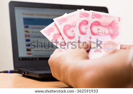 Hand holding China Yuan  in office with computer screen showing foreign exchange table in background