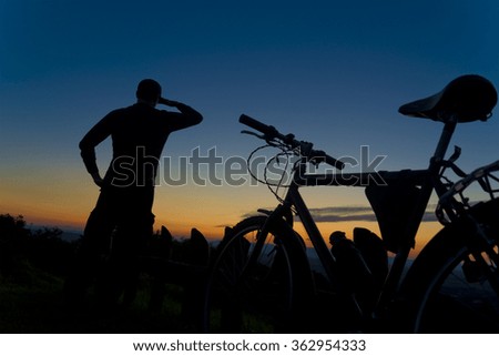 Silhouette of a cyclist on top of mountain watching landscape with Sunset. Male standing next to mountain bike bicycle. One hand is above eyes forming shade while other hand is resting on hip.