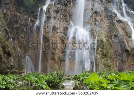 Photo of beautiful waterfalls cascades falling down from mountain wall rock surrounded by picturesque green rich foliage on natural landscape background, horizontal picture