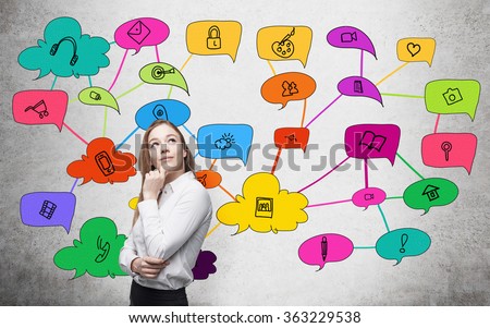 Young woman looking upwards with hand on her chin thinking about functions of social networks depicted as coloured icons around her. Concrete background. Concept of social networks.