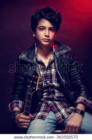 Portrait of handsome teen guitarist posing over dark red background, wearing trendy shirt and leather coat, cool teen's hobby