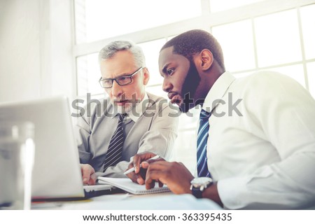 Photo of aged businessman and his colleague. Businessmen working in office with big window. Men using laptop