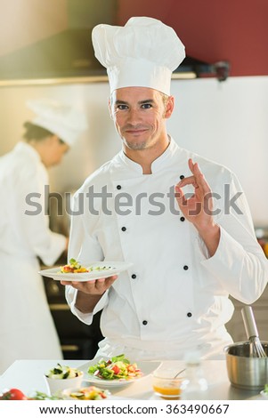 A man chef in his fifties is standing in a professional kitchen presenting a plateful with fine food. He is looking at camera doing the ok sign, wearing white chef clothes and hat. Blurred background.