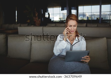 Woman owner takes order from client via mobile phone for conduct of corporate party while sitting in her restaurant, young female looking at digital tablet screen while talking via cell telephone 