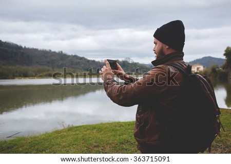 Young hipster guy taking photo on his mobile phone camera of a beautiful landscape while standing near lake, bearded man shoots video on cell telephone during amazing travel through the countryside