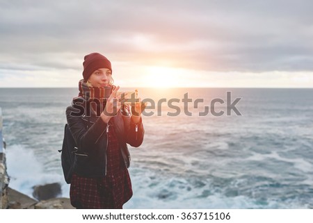 Trendy woman photographing nature with mobile phone camera while standing against sunset and blue sea background, young female taking pictures of a beautiful ocean landscape on cell telephone