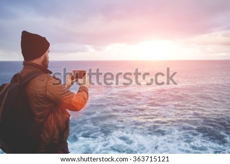Back view of a young man tourist taking photo with cell telephone digital camera while standing in front sea with waves, hipster guy with trendy look shoots video with ocean landscape on mobile phone 