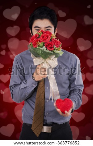 Picture of young Chinese man holding a bouquet of flower and a gift to give a surprise at his girlfriend, shot with heart background