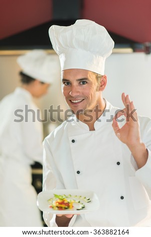 A man chef in his fifties is standing in a professional kitchen presenting a plateful with fine food. He is looking at camera doing the ok sign, wearing white chef clothes and hat. Blurred background.