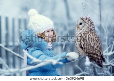 Cute little girl in warm clothing looking at an eagle Owl sitting on a branch covered with frost on a cold winter day. Children and nature.