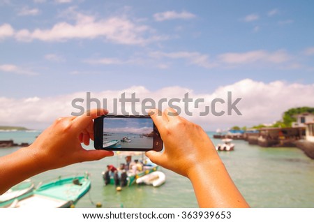 Hands holding mobile phone taking picture of beautiful island bay area on Galapagos islands with nice weather and small boats
