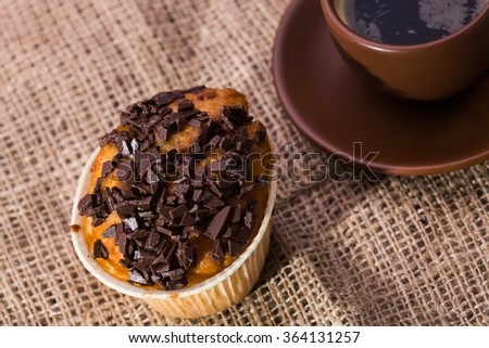 Tasty homemade warm muffin with chocolate pieces and beautiful brown cup of black fragrant coffee on plate lunch time closeup on sackcloth background top view, horizontal picture