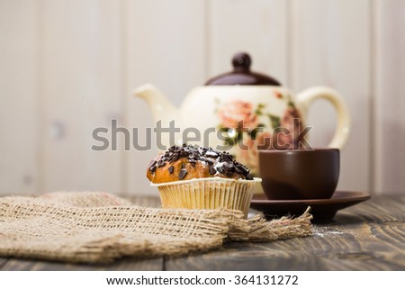 Appetizing breakfast fresh cupcake with aromatic black coffee in brown cup and beautiful teapot near sackcloth napkin on wooden table light background indoor closeup, horizontal picture