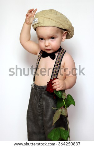 Little boy in cap, suspenders and bow-tie with one hand on a cap and holding a red rose in another one greeting somebody, isolated portrait on white background, romantic and love concept
