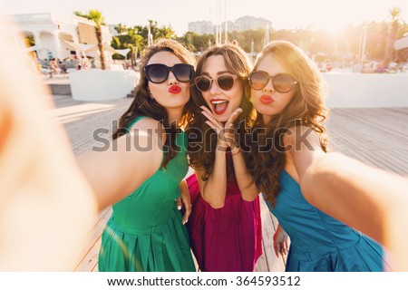Summer outdoor portrait of three friends fun girls  taking photos with a smartphone at bright sunset. Group of happy women  taking self-portrait on their travel vacations. Wearing colorful dress. Royalty-Free Stock Photo #364593512