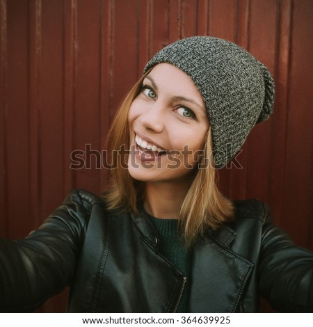Trendy beautiful cool hipster blond girl wearing a gray hat, black leather jacket taking selfie. Smiling girl. Urban style. Edited with filter.