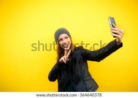 Trendy beautiful cool hipster blond girl wearing a gray hat and black leather jacket taking selfie with mobile phone against a yellow wall. Making peace hand gesture and smiling. 