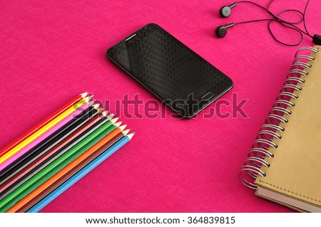 A slim phone with earphones displayed with a note book and coloring pencils on a pink background