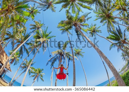 Beautiful girl fly high with fun in blue sky on rope swing among coconut palms on sea beach in tropical island. Healthy lifestyle, people activity and relaxation on summer family vacation with child.