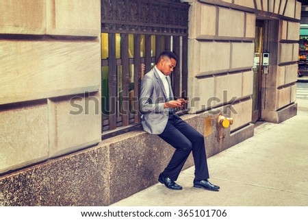 African American man reading, texting on cell phone on street, traveling, working in New York, wearing gray blazer, black pants, leather shoes, sitting on vintage window frame. Instagram filtered look