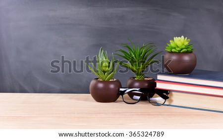 Classroom desktop displaying textbooks, plants, reading glasses with blank blackboard in background. Selective focus on front part of desk objects.