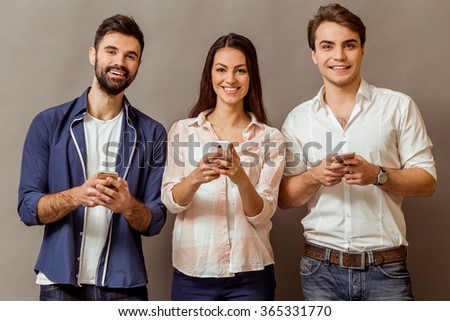 Group of attractive young people holding a smartphone, looking at camera, smiling, on a gray background