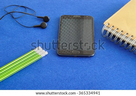 A slim phone with earphones displayed with a note book and writing pencils on a blue background