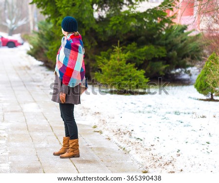 Girl walking by sidewalk at winter, dressed in warm clothes and blanket with grid