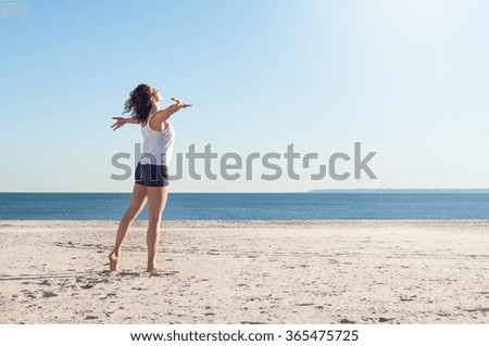 Happy young woman with arms outstretched at beach on a bright morning. Beautiful young woman with arms stretched out standing on a beach. Happy young woman feelling good and relaxing at the beach.  Royalty-Free Stock Photo #365475725