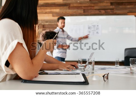 Closeup image of young woman sitting in a meeting in boardroom with man giving presentation in background. Focus on hands of female executive.
