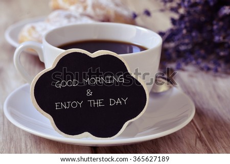 the text good morning and enjoy the day written in a thought bubble-shaped blackboard placed in a cup of coffee, with some pastries in the background in a set table for breakfast