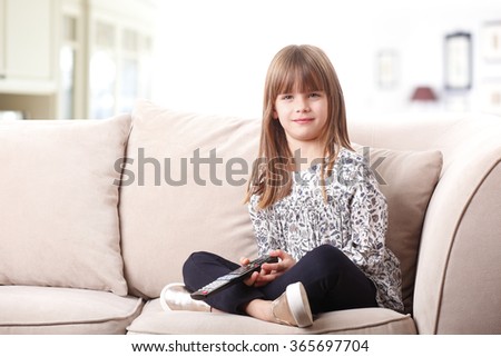 Portrait of adorable little girl sitting on couch and holding in her hand a remote control while watching cartoon at tv.