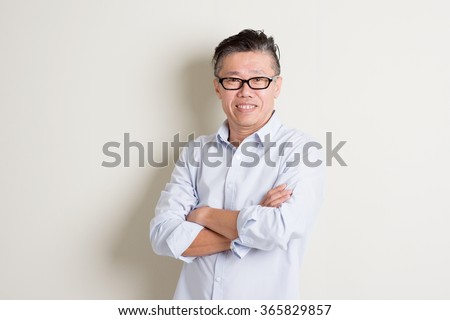 Portrait of happy single mature 50s Asian man in casual business arms crossed smiling and standing over plain background with shadow.