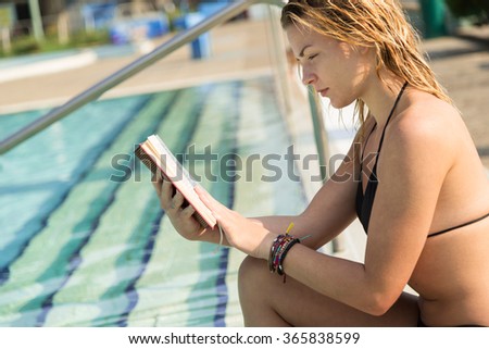 Young, beautiful blond girl sitting next to a swimming pool and reading a book