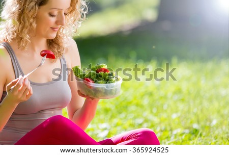 Young slim woman holding a bowl of fresh salad ofter working out outdoors in the park. Eating on fresh air, sitting on the grass and enjoying early morning. Healthy lifestyle.