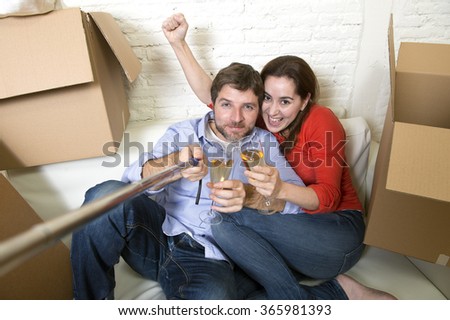 young happy American couple sitting on couch unpacking boxes together celebrating with champagne toast moving in a new house or apartment flat taking selfie photo in real estate concept