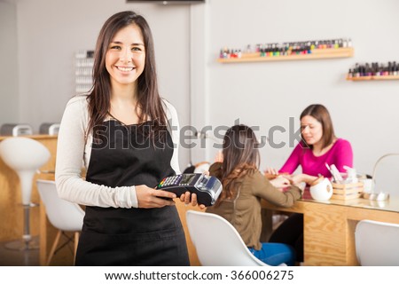 Portrait of a cute young Latin woman holding a credit card terminal at a nail salon and smiling