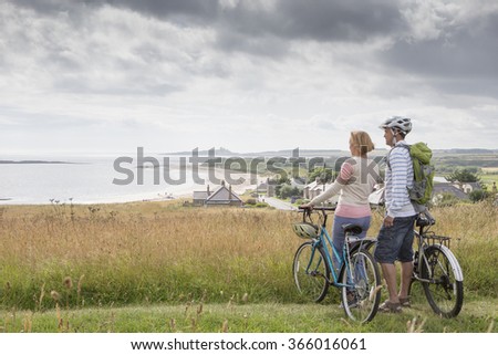A couple are enjoying the view on the sand dunes. They are holding their bikes, looking at the beach.
