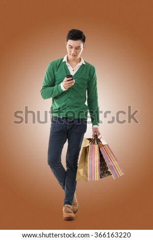 Asian young man holding shopping bags and using cellphone, full length portrait isolated.