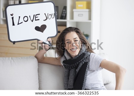 Beautiful woman sitting in living room and holding a white board with written word "I love you"