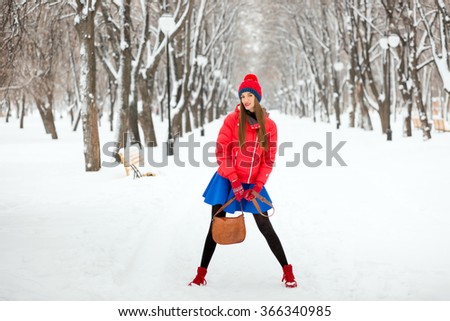 Beautiful winter portrait of young woman in the winter snowy scenery. The girl in a red hat, a red jacket and blue skirt, posing in a park in winter.