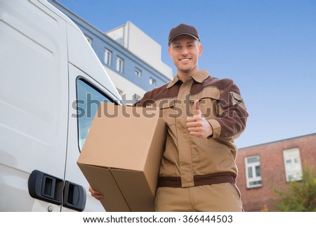Low angle portrait of delivery man carrying cardboard box while gesturing thumbs up by truck