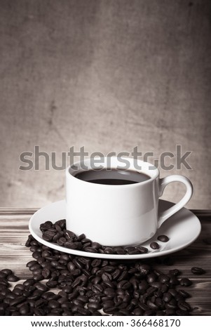 Coffee beans and coffee in white cup on wooden table opposite a defocused burlap background. Toned.